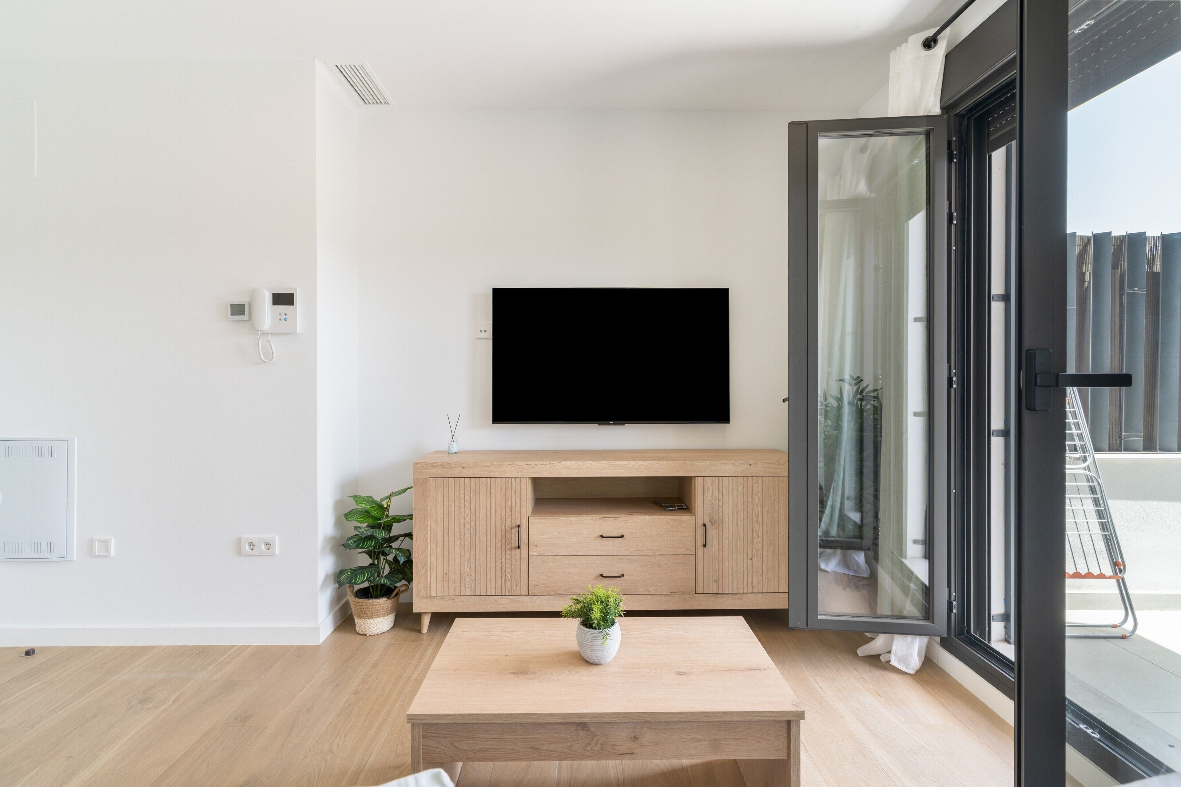 Un salón moderno con televisor de pantalla plana, muebles de madera, una mesita con una planta y una puerta corrediza de vidrio que da a un espacio exterior.