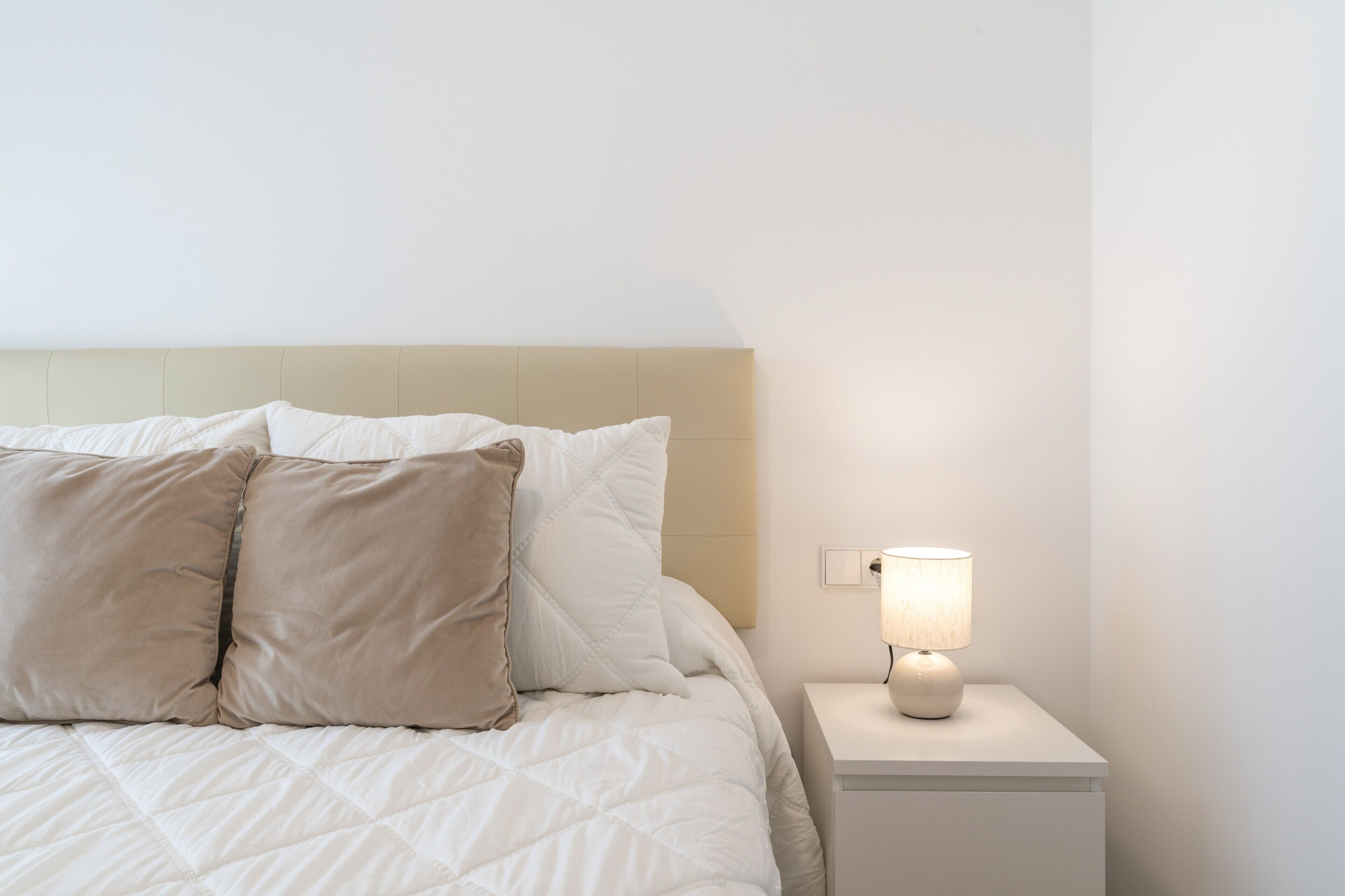 Una cama bien hecha con edredón blanco acolchado y dos almohadas color beige, una lámpara de noche en una mesita blanca, y una pared blanca lisa.