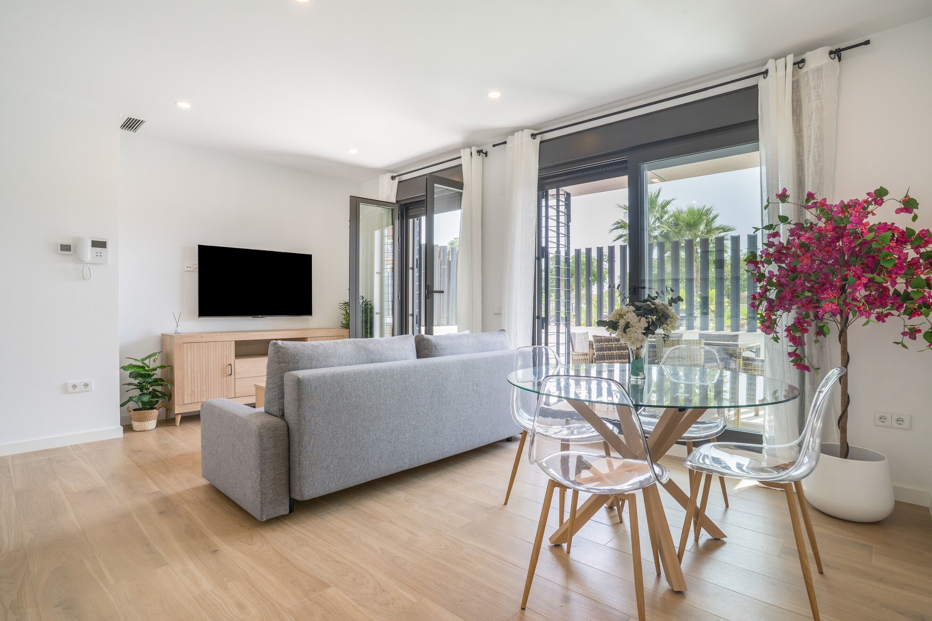 Una sala de estar moderna con un sofá gris, una mesa de comedor de cristal con sillas, un televisor de pantalla plana y un ventanal con cortinas.
