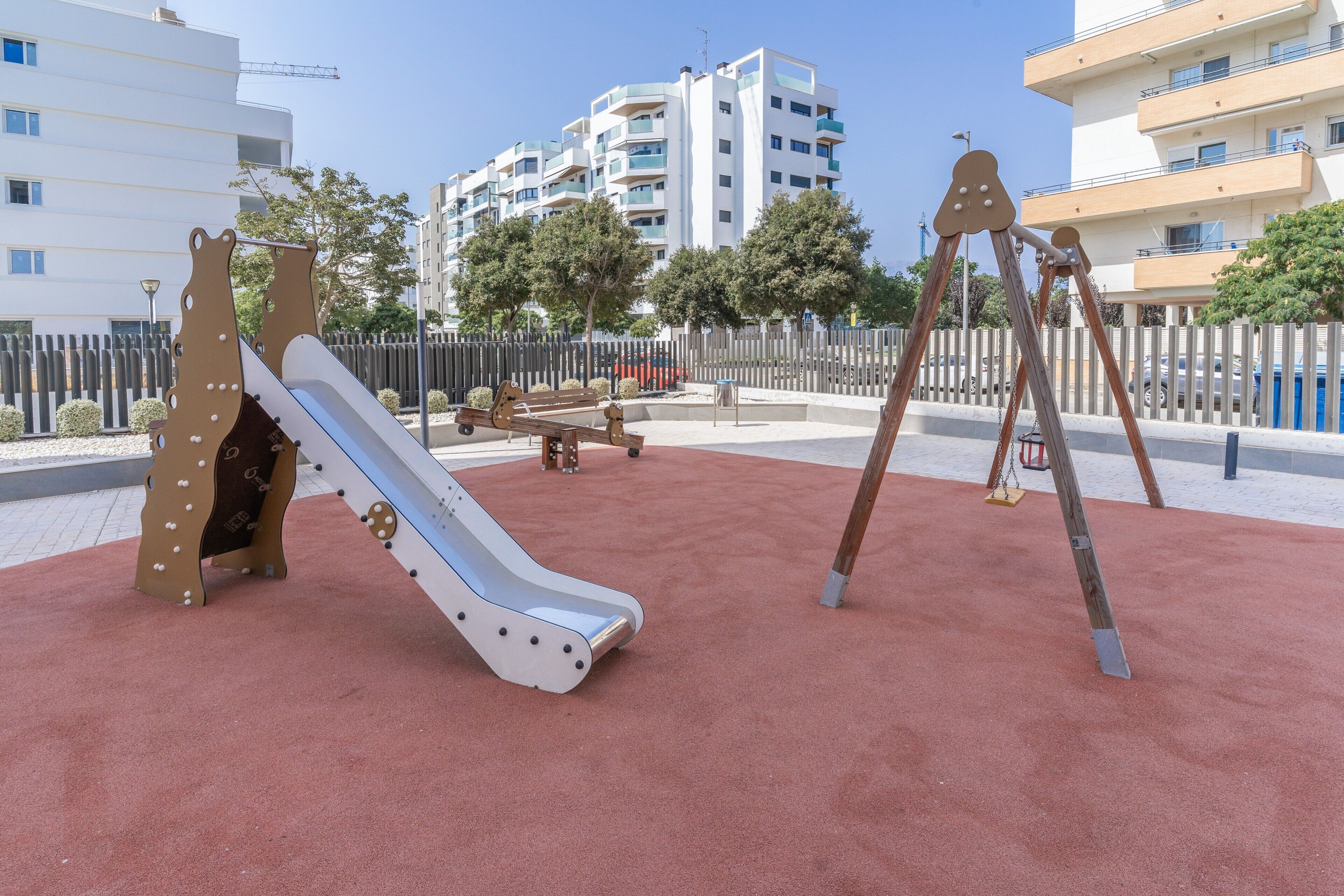 Un parque infantil con un tobogán y columpios, rodeado por una valla, con edificios al fondo.