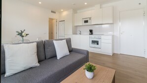 Een moderne woonkamer met een grijze bank, een bijzettafeltje met een plant en een keuken met witte kasten en apparatuur.