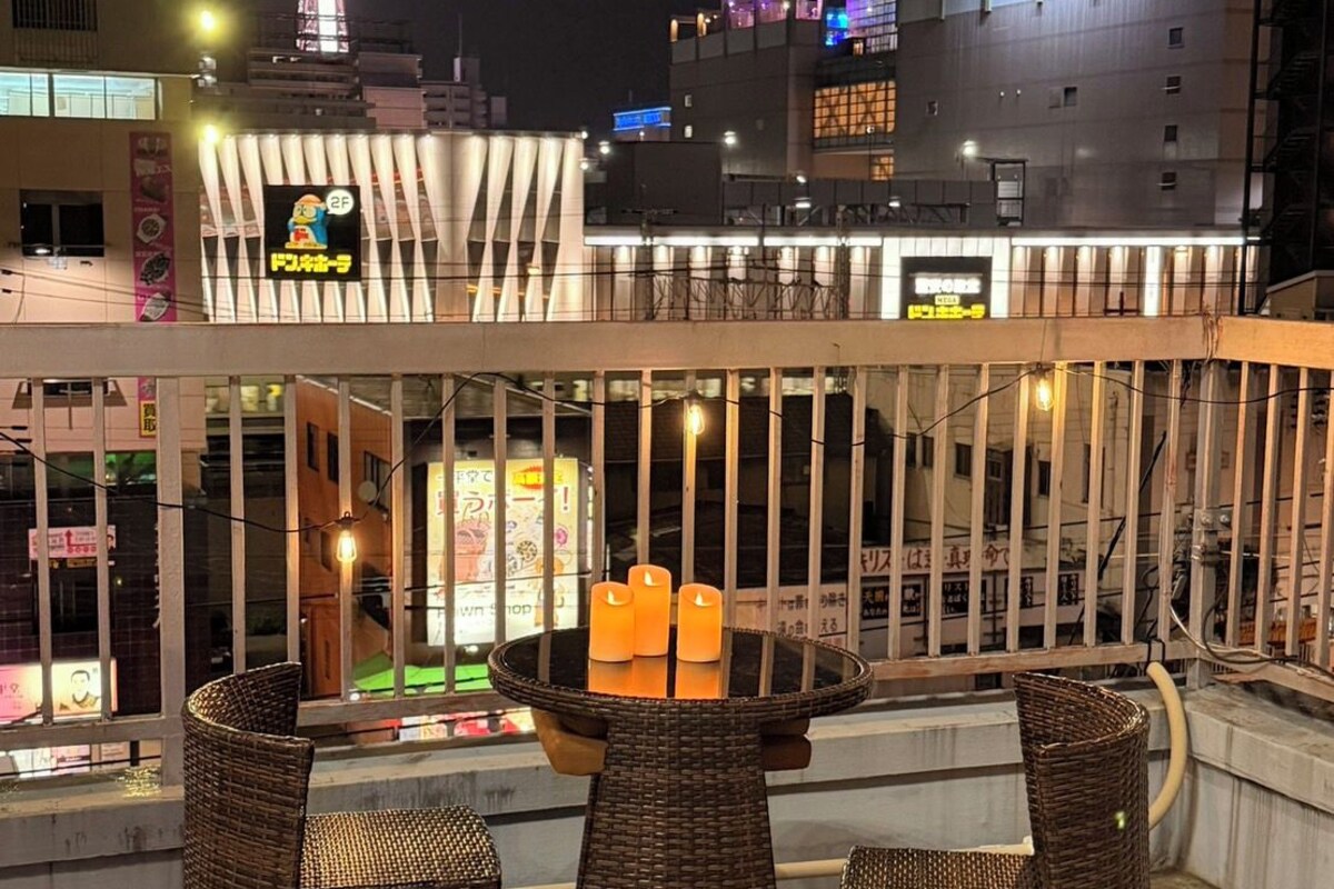 Un área de estar al aire libre con mobiliario de mimbre y una mesa con velas, con vistas a un paisaje urbano con edificios iluminados y una torre.