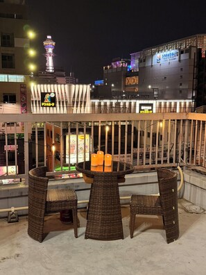 Un área de estar al aire libre con mobiliario de mimbre y una mesa con velas, con vistas a un paisaje urbano con edificios iluminados y una torre.