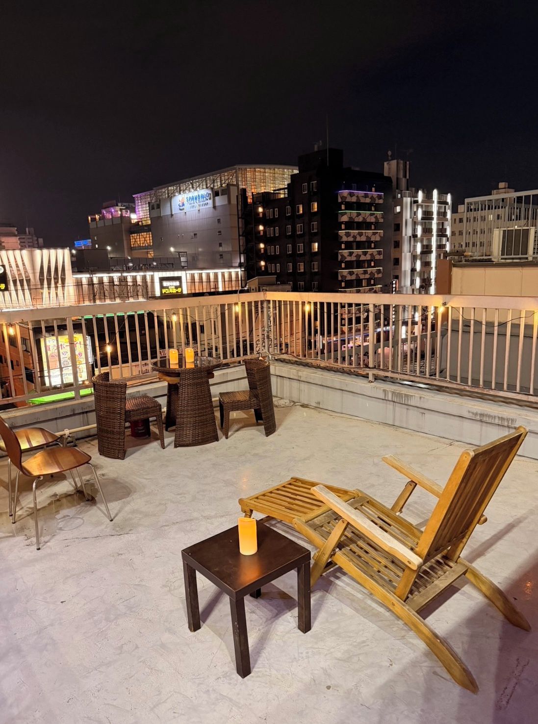 Una terraza en la azotea con mobiliario de mimbre y una mesa, con vistas a un paisaje urbano nocturno.