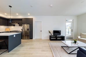 A modern kitchen with a central island, a dining area with a coffee table, and a living room with a sofa and a TV. - Brand New Luxury 2 Bedroom Surrey/White Rock (Surrey)