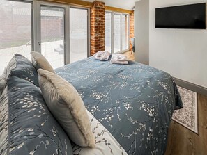 A bedroom with a large bed, a flat-screen TV mounted on the wall, and a view through the window. - Rose Blossom Cottage (Benniworth, near Market Rasen)