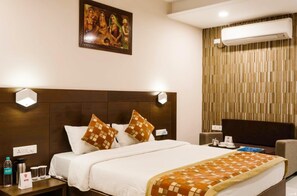 A hotel room with a large bed, a headboard with wall-mounted lights, a painting above the bed, a wall-mounted air conditioner, and a side table with a water bottle and a sign. - Hotel Raj Ganga In Haridwar (Haridwar)