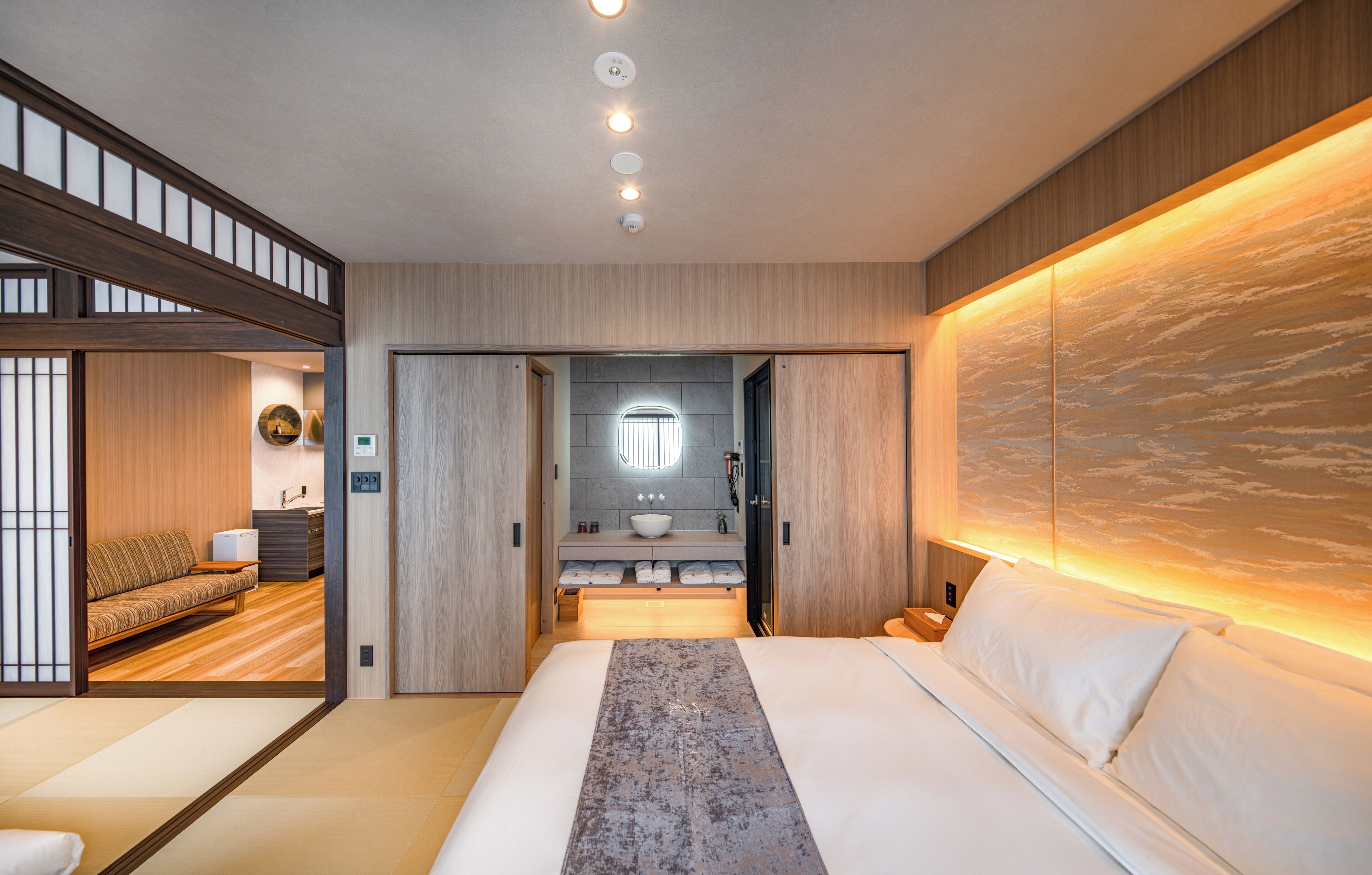 A modern hotel room with a large bed, a sitting area with a sofa, a bathroom with a large mirror, and a view of the sky through a window.