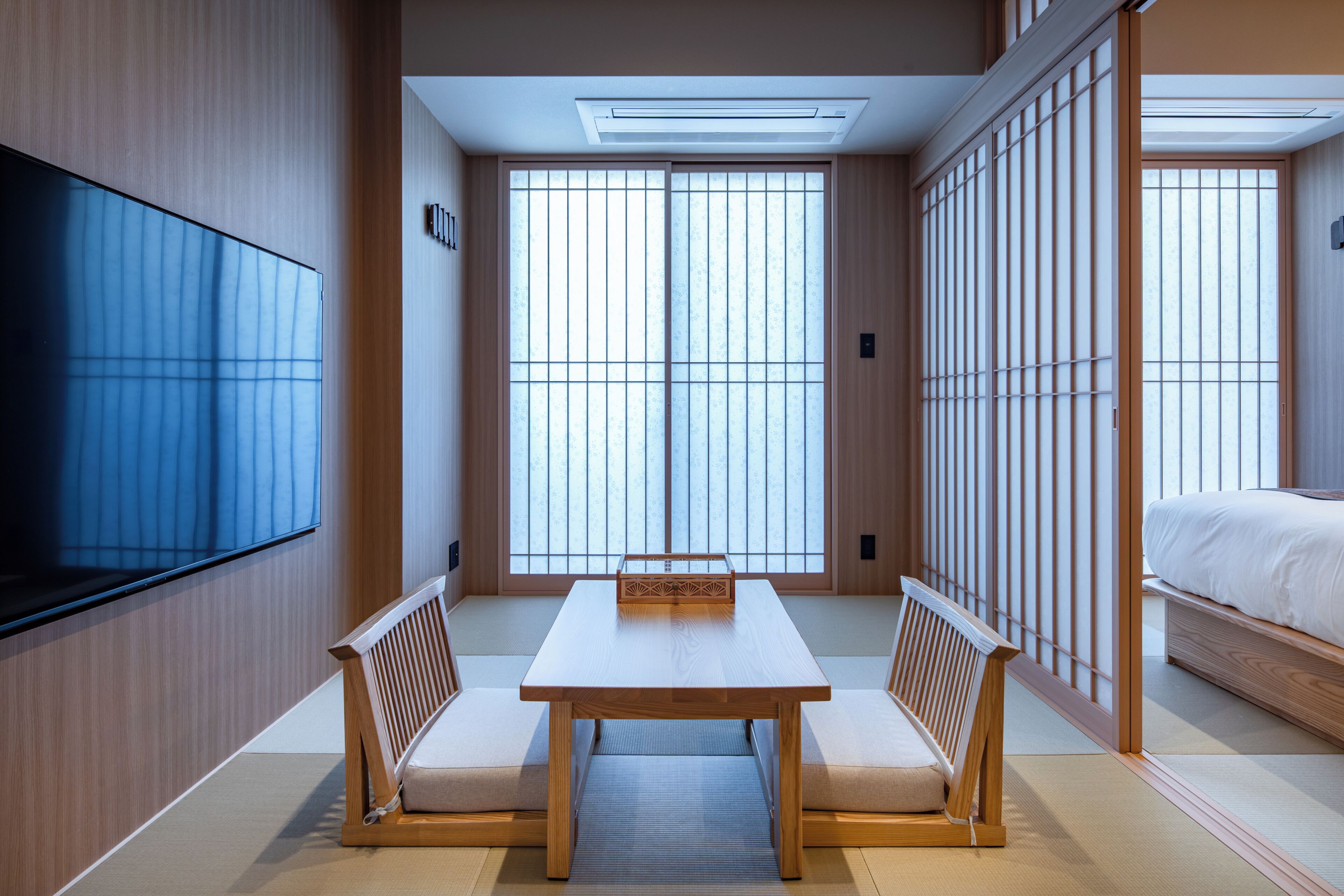 A traditional Japanese-style room with tatami flooring, a low table with cushions, a television, and sliding doors.