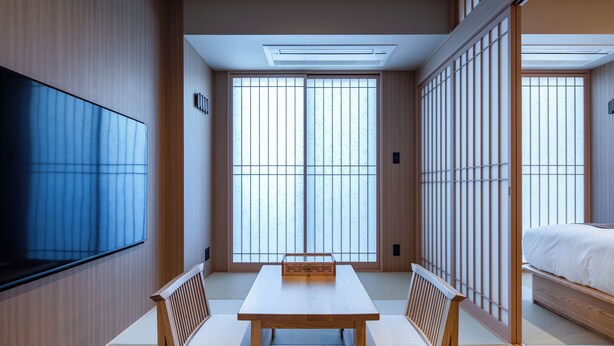 Una habitación tradicional japonesa con suelo de tatami, una mesa baja con cojines, un televisor y puertas correderas.