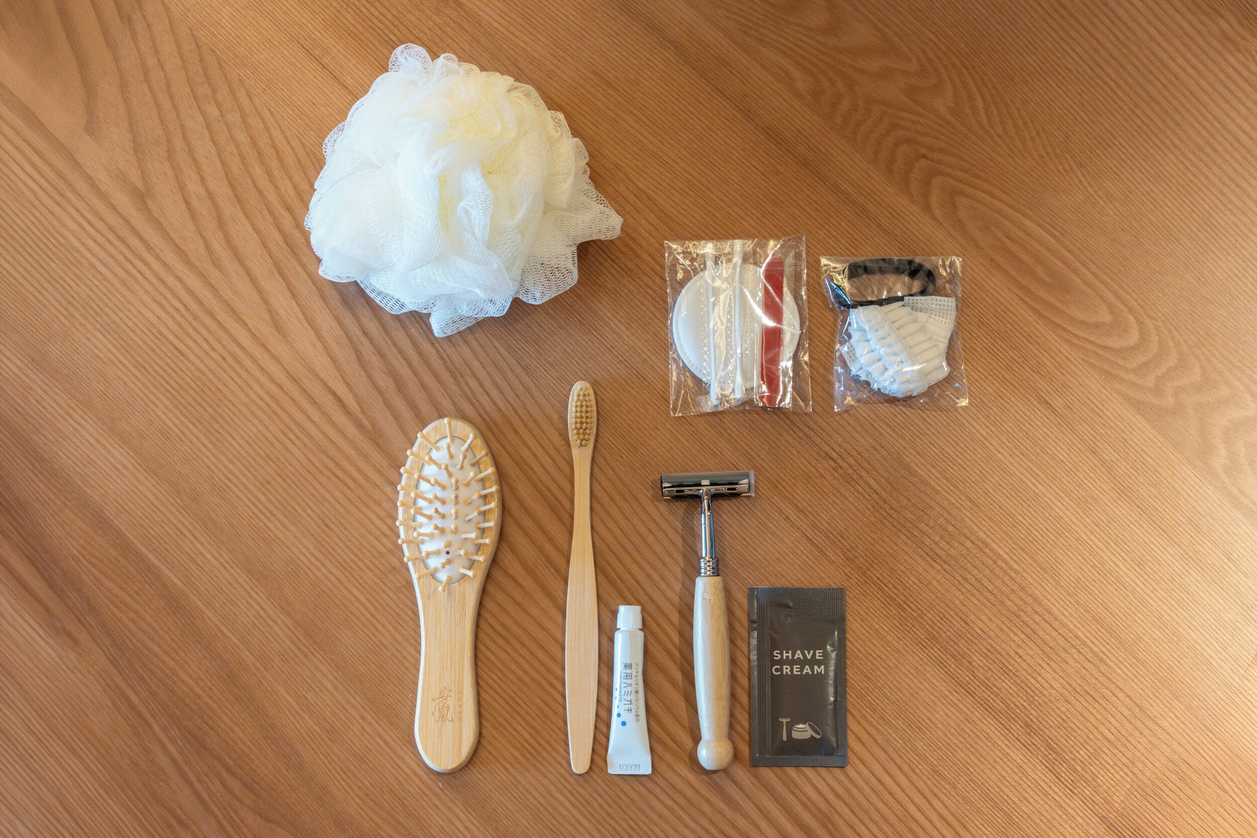 A wooden surface with a white bath sponge, a wooden brush, a razor, a shaving cream tube, and a razor blade set.