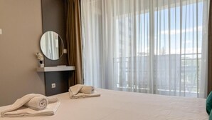 Quarto de hotel com cama, espelho redondo e janela com cortinas translúcidas.