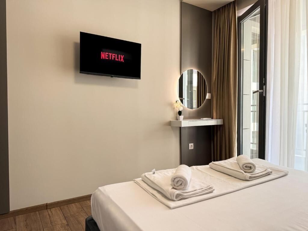 Habitación de hotel con cama, televisor montado en la pared mostrando 'NETFLIX', espejo redondo y ventana con cortinas.