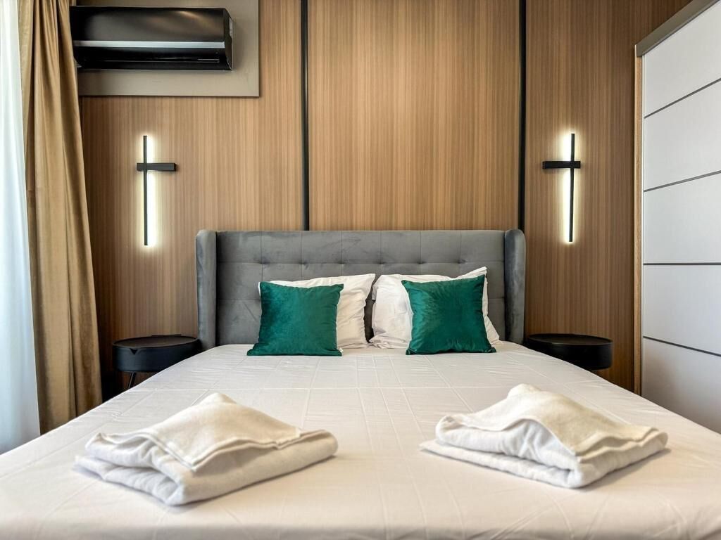 A hotel room with a large bed, two bedside tables, a wall-mounted air conditioning unit, and a cross-shaped light fixture.