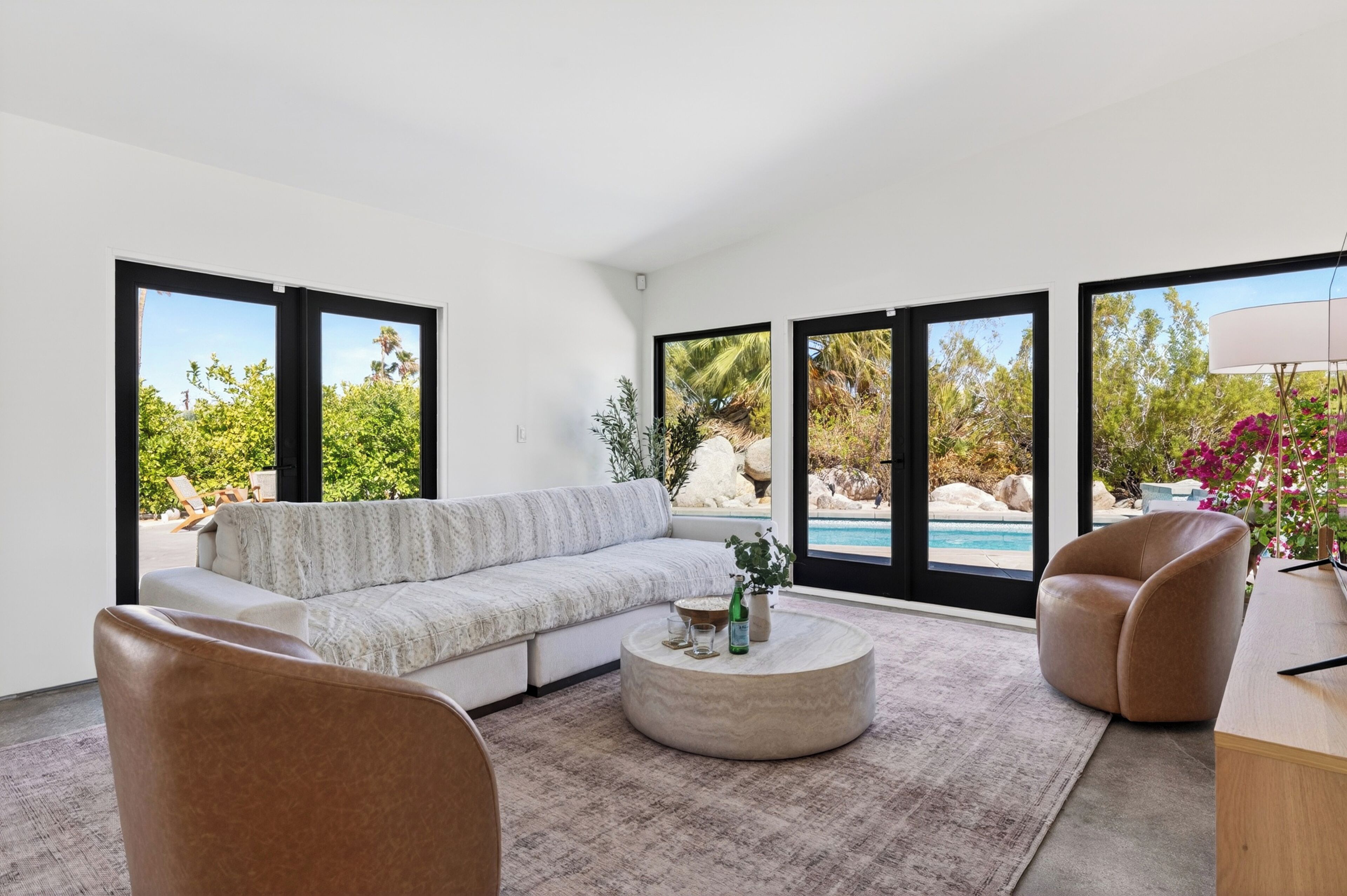 A modern living room with a sectional sofa, a coffee table, and two armchairs. Large windows provide views of a pool and outdoor greenery.