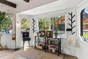 A room with white paneled walls, a black chair, a table with books and a plant, and a view of the outside. - Hot Tub + Game Room + Firepit • Near Bear Mountain + Lake | Balboa Big Bear (Big Bear Lake)