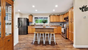 Una cocina moderna con armarios de madera, un refrigerador de acero inoxidable, una isla central con taburetes y un ventanal que proporciona luz natural.