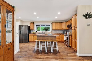 A modern kitchen with wooden cabinets, a stainless steel refrigerator, a central island with stools, and a large window providing natural light. - Firepit + Grill + Chef Kitchen | Creek + Waterfalls | Ohana Nature Escape (Catskill)