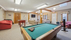 A pool table with a cue ball, a red sofa, and a television in a room with wooden beams and a carpeted floor. - 2 Townhomes + Indoor Pool + Game Room | Gore Mtn | The Summit at Gore Mtn (North Creek)