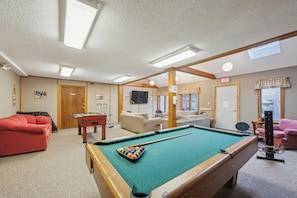 A pool table with a cue ball, a red sofa, and a television in a room with wooden beams and a carpeted floor. - 2 Townhomes + Indoor Pool + Game Room | Gore Mtn | The Summit at Gore Mtn (North Creek)