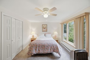 Une chambre à coucher avec un lit, deux lampes de chevet, un ventilateur de plafond, une fenêtre donnant sur un beau paysage et un radiateur.