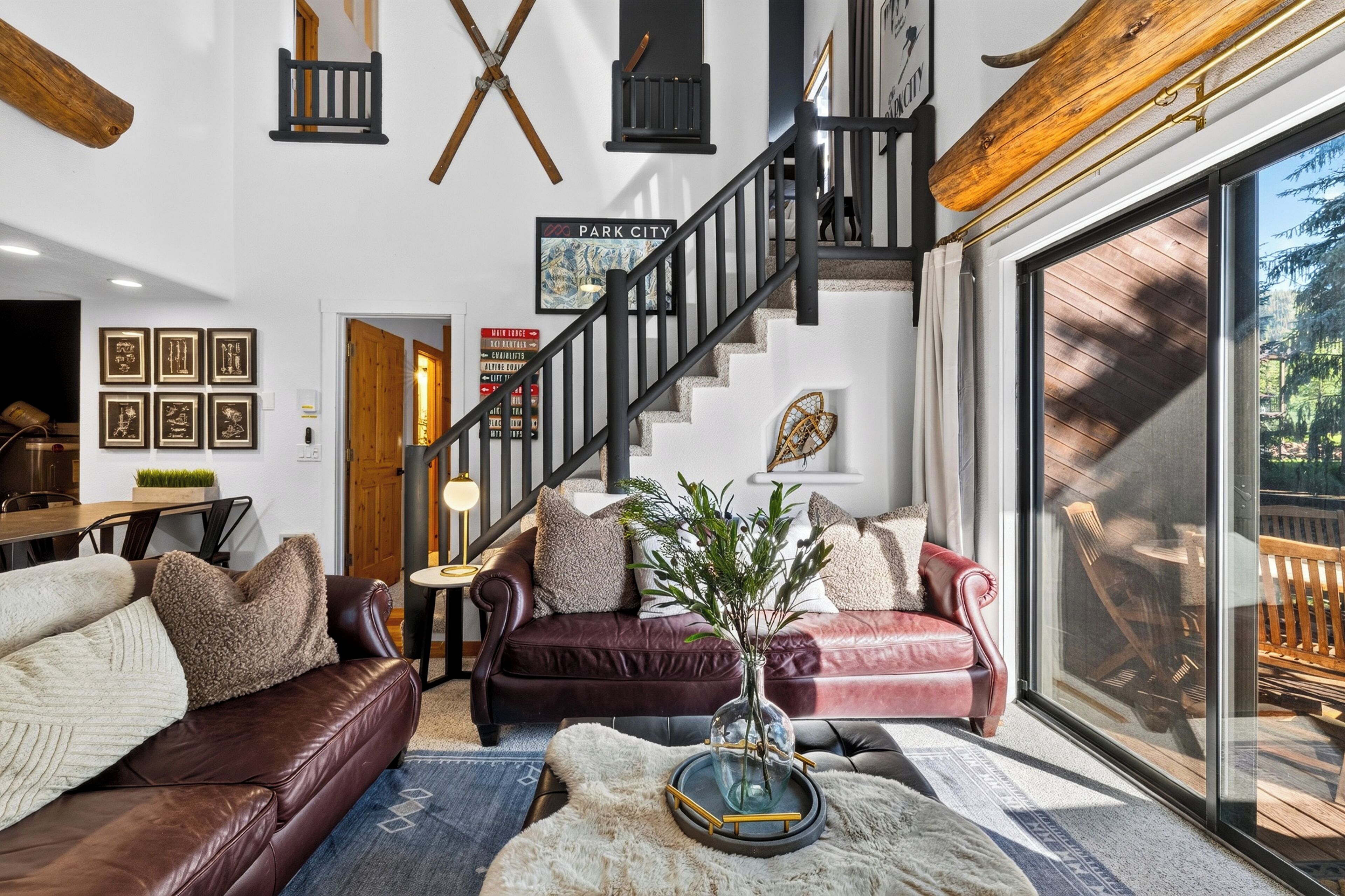 A spacious living room with a staircase, leather sofas, a coffee table with a vase, and a large window offering a view of the outdoors.