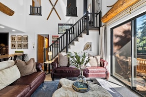 Uma sala de estar ampla com escada, sofás de couro, uma mesa de centro com um vaso e uma grande janela com vista para o exterior.