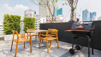 Een terras buiten met een tafel, stoelen, een barbecue en een stadsachtergrond.