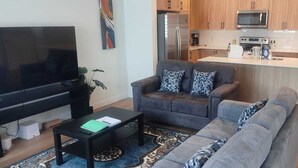 A living room with a flat-screen TV, grey sofa, patterned cushions, and a coffee table. A kitchen with wooden cabinets and modern appliances is visible in the background. - Spacious 3BR Townhouse in SE Calgary – Perfect for Families & Groups (Calgary)