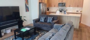 Ein Wohnzimmer mit Flachbildfernseher, grauem Sofa, gemusterten Kissen und einem Couchtisch. Im Hintergrund ist eine Küche mit Holzschränken und modernen Geräten zu sehen.