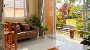 A modern living room with a sofa, dining table, and chairs, a sliding glass door leading to a garden with a wooden house and palm trees.