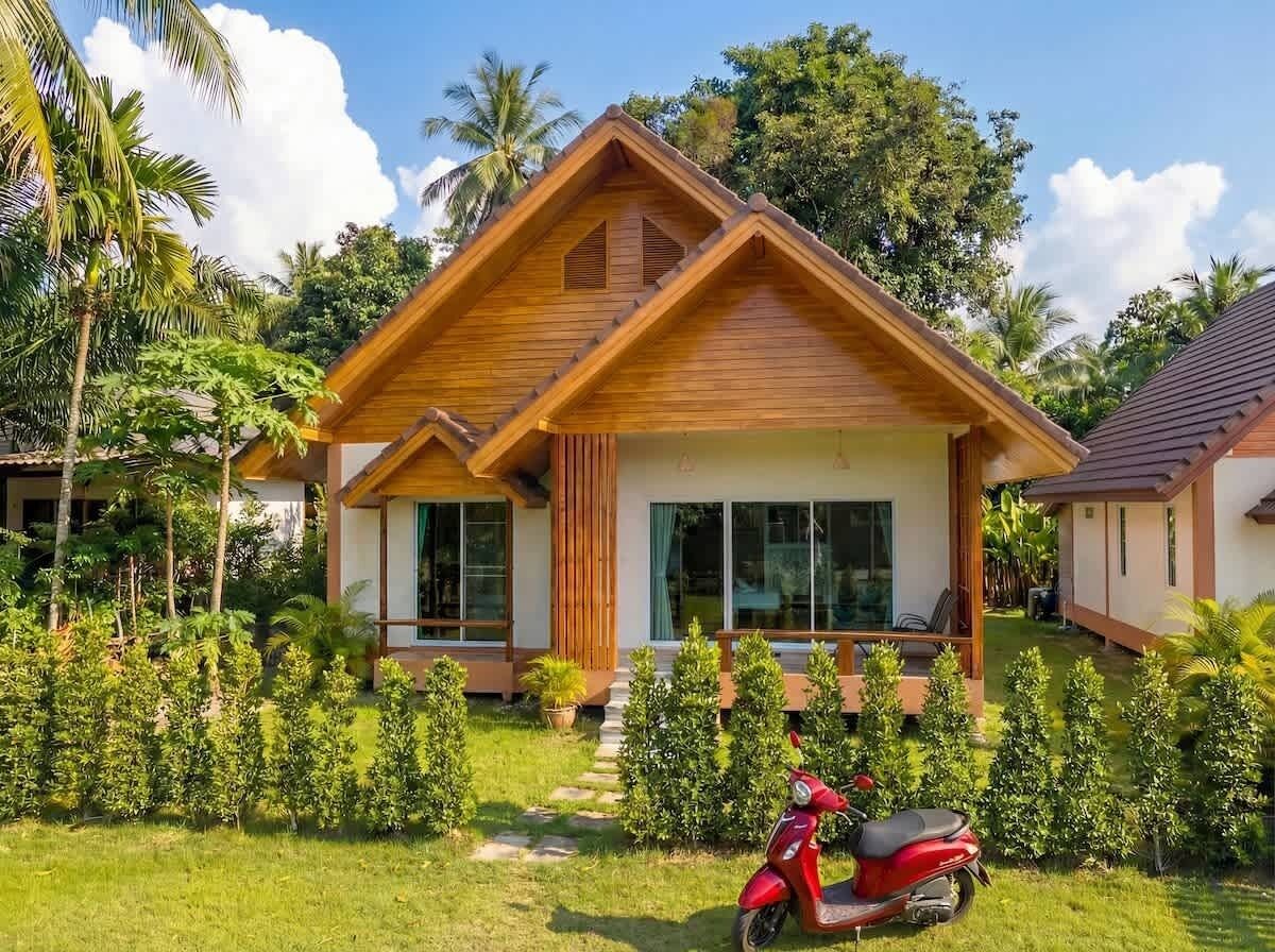 A wooden house with a sloped roof, a red scooter parked in front, and a well-maintained lawn with small trees.