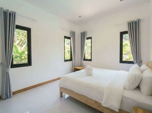 A bedroom with a large bed, two windows with curtains, and a view of greenery outside.