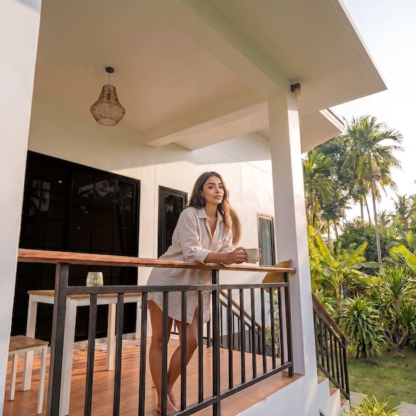 A person on a balcony with a railing, a hanging light fixture, and a view of greenery and palm trees.