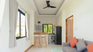 Uma sala de estar moderna com uma mesa de centro de vidro, um sofá cinza com almofadas laranja, um ventilador de teto e uma área de cozinha com geladeira e banquetas de bar.