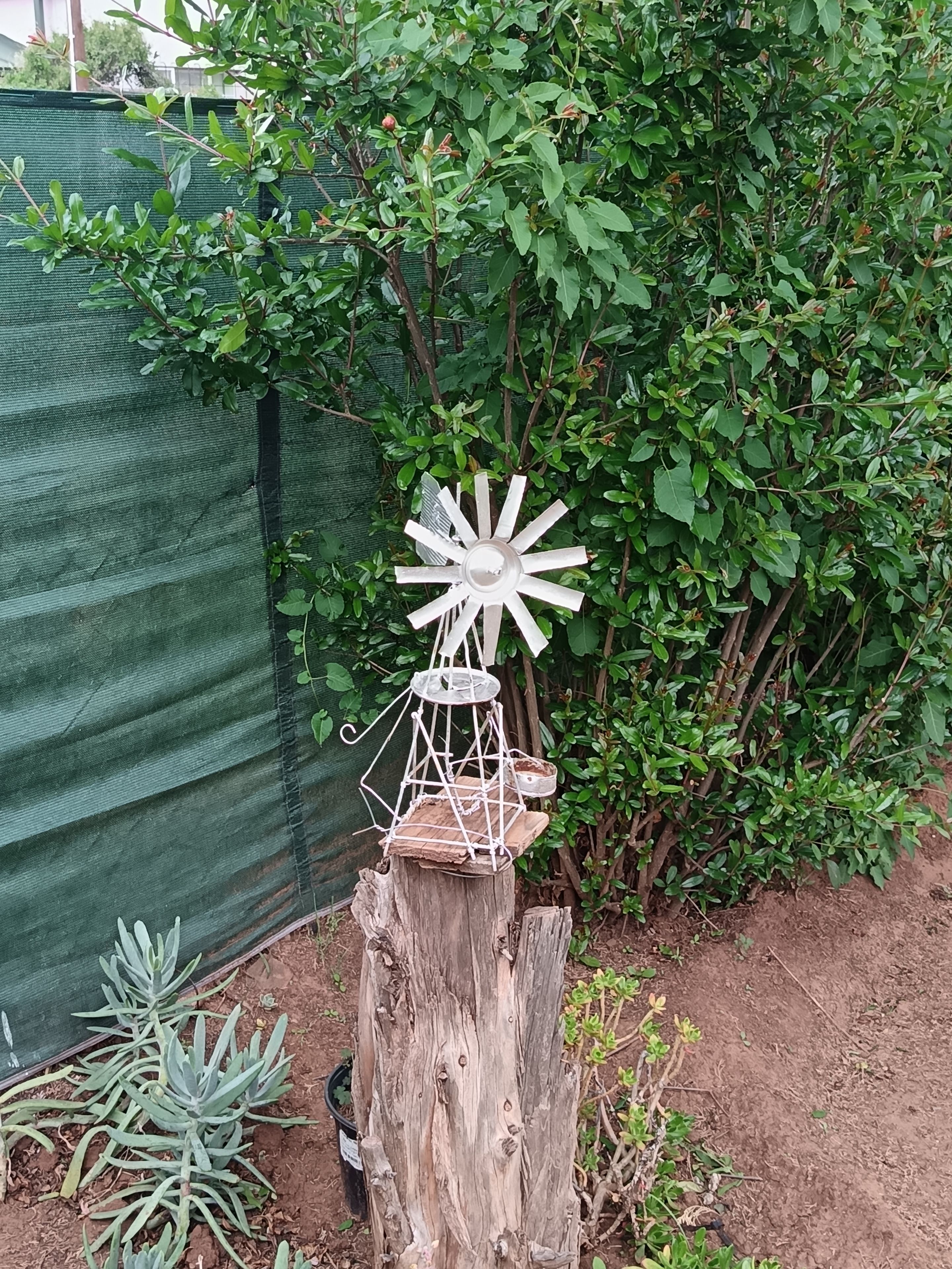 Un poteau en bois rustique, surmonté d’un moulin à vent blanc en guise d’ornement et d’une petite table, le tout entouré de verdure et délimité par une clôture en treillis vert.