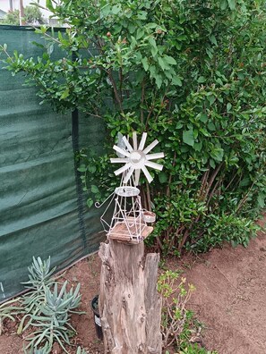 Un poste de madera rústico con un adorno de molino blanco y una pequeña mesa en la parte superior, rodeado de follaje y una cerca de malla verde.