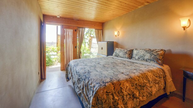 A bedroom with a wooden ceiling, a bed with patterned bedding, a nightstand, and a wall-mounted light fixture. - Serene, Family-Sized Off Grid Earthship w/ Big Mtn (Tres Piedras)