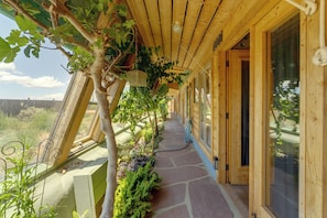 A covered wooden walkway with a stone floor, surrounded by various plants and a view of the landscape. - Serene, Family-Sized Off Grid Earthship w/ Big Mtn (Tres Piedras)