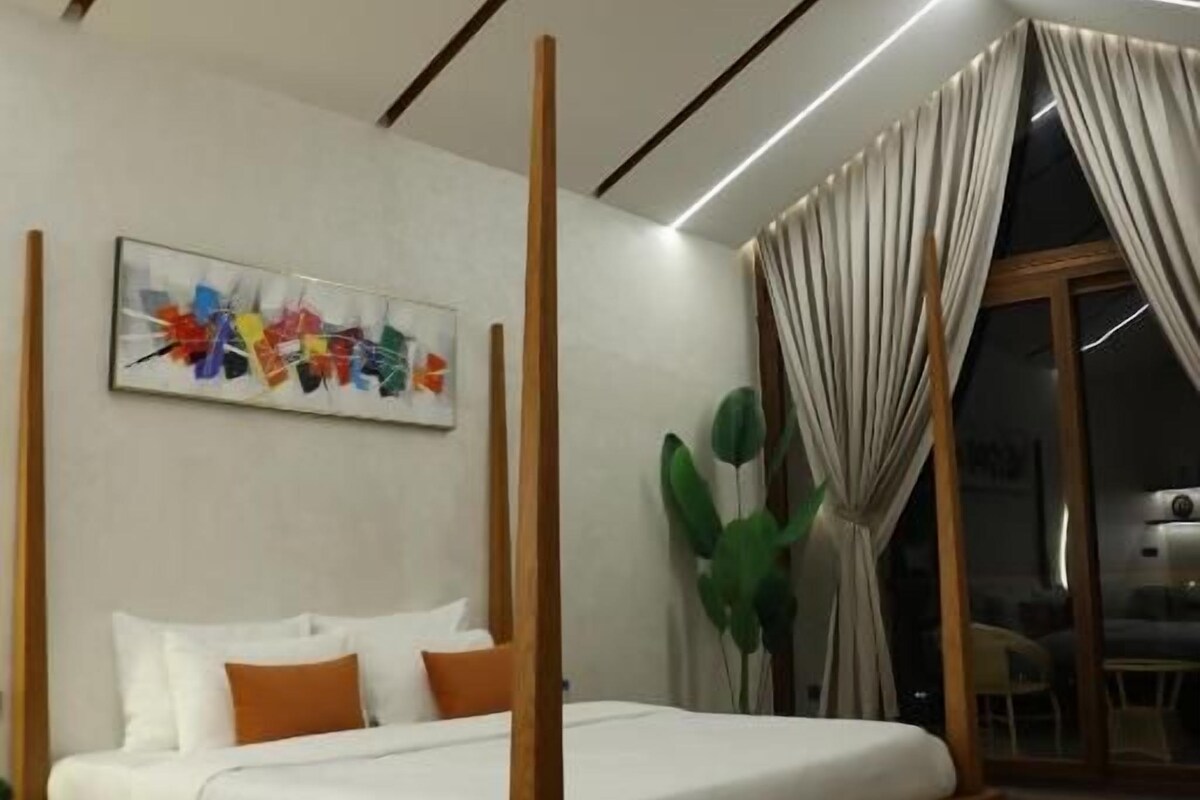 A modern bedroom with a four-poster bed, a large abstract painting, and a view of the outdoors through a glass door.