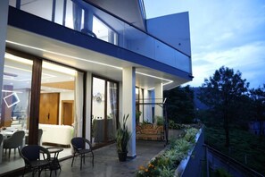 Exterior moderno de hotel com fachada de vidro, área de estar externa e varanda com vista para um jardim paisagístico.