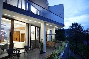 A modern hotel exterior with a glass facade, outdoor seating area, and a balcony overlooking a landscaped garden.