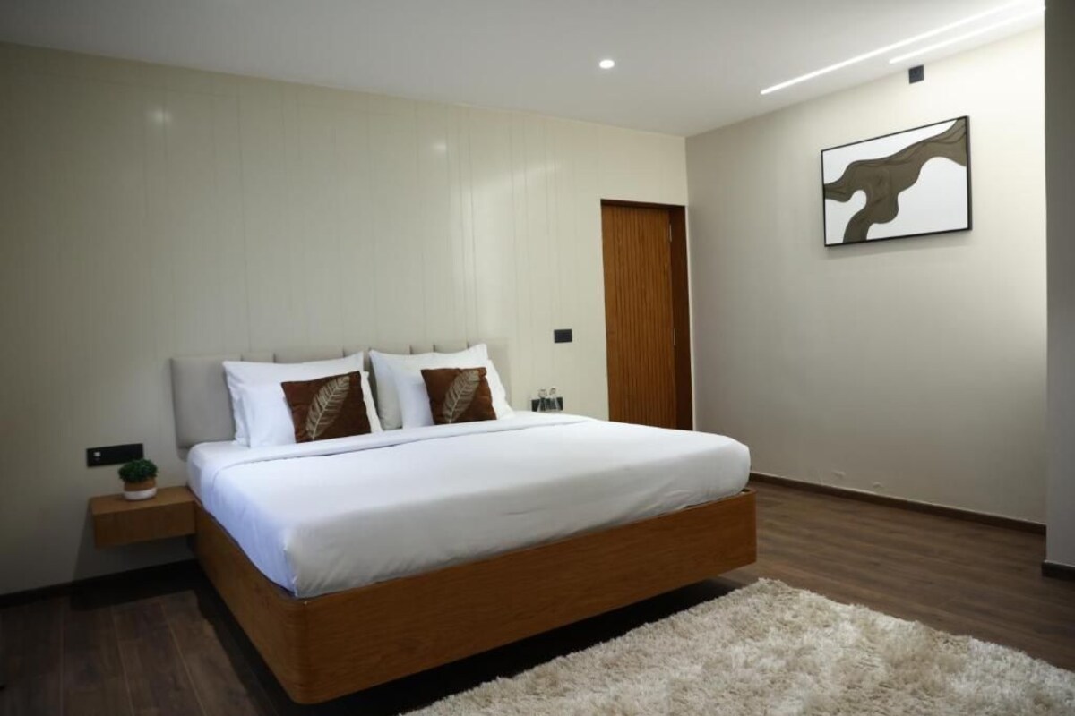 A modern bedroom with a large bed, a wooden headboard, a fluffy rug, a nightstand with a plant, and a framed artwork on the wall.