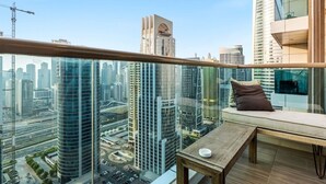 A high-rise building with a balcony featuring a wooden table and a cushioned seat, overlooking a cityscape with modern skyscrapers. - Relaxing City Escape in JLT (Dubai)