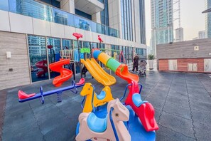 An outdoor playground with slides and swings, located in front of a modern building with large windows. - Relaxing City Escape in JLT (Dubai)