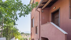 Un edificio con fachada roja, una ventana con persianas y un balcón con plantas en macetas.