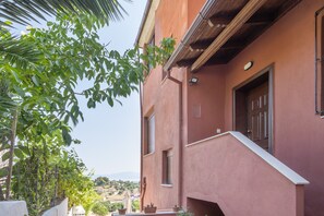 A building with a red facade, a window with shutters, and a balcony with potted plants.
