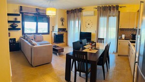 Una sala de estar con un sofá, una mesa de centro y un televisor. Hay un comedor con una mesa y sillas. La habitación tiene una chimenea y un reloj en la pared.