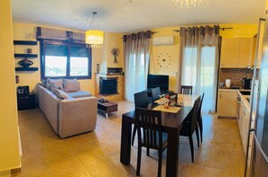 A living room with a sofa, a coffee table, and a TV. There's a dining area with a table and chairs. The room has a fireplace and a clock on the wall.