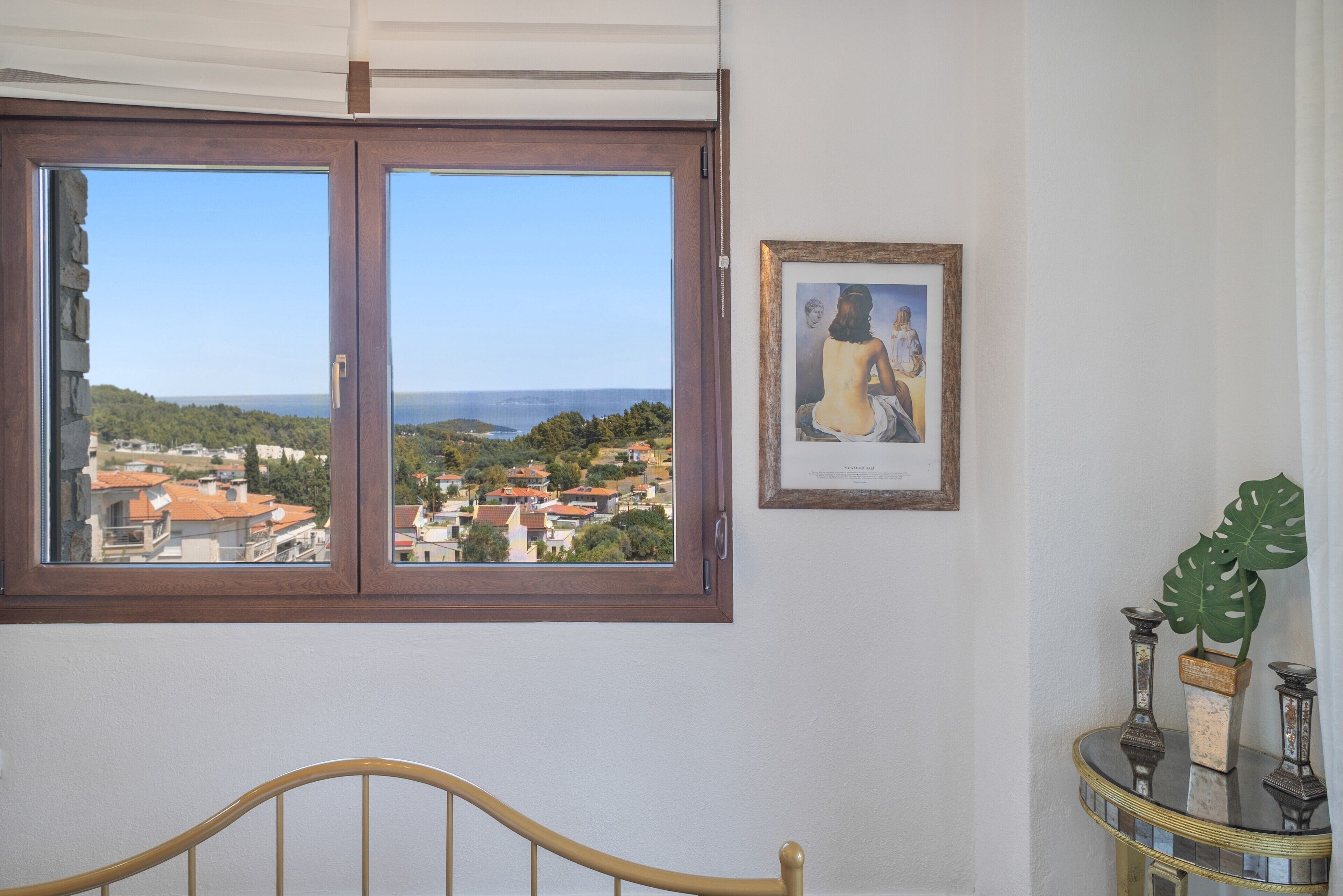 Ein Zimmer mit großem Fenster mit Blick auf eine Küstenstadt, einem gerahmten Kunstwerk an der Wand und einem kleinen Tisch mit Dekoration.